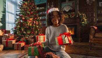 Merry Christmas. African American woman packing wrapping gift box near Christmas tree. Girl in living room with Christmas tree and fireplace opening gift box. Christmas eve at home.