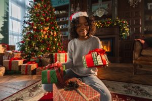 Merry Christmas. African American woman packing wrapping gift box near Christmas tree. Girl in living room with Christmas tree and fireplace opening gift box. Christmas eve at home.