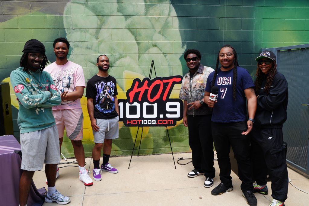 A group of 6 people posing in front of a "HOT 100.9" sign, wearing casual clothing and hats.