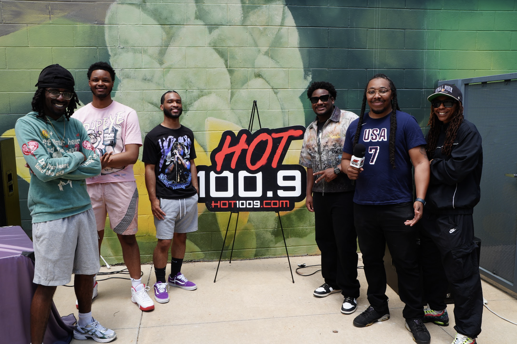 A group of six people posing in front of a "HOT 100.9" sign, wearing casual clothing and accessories.