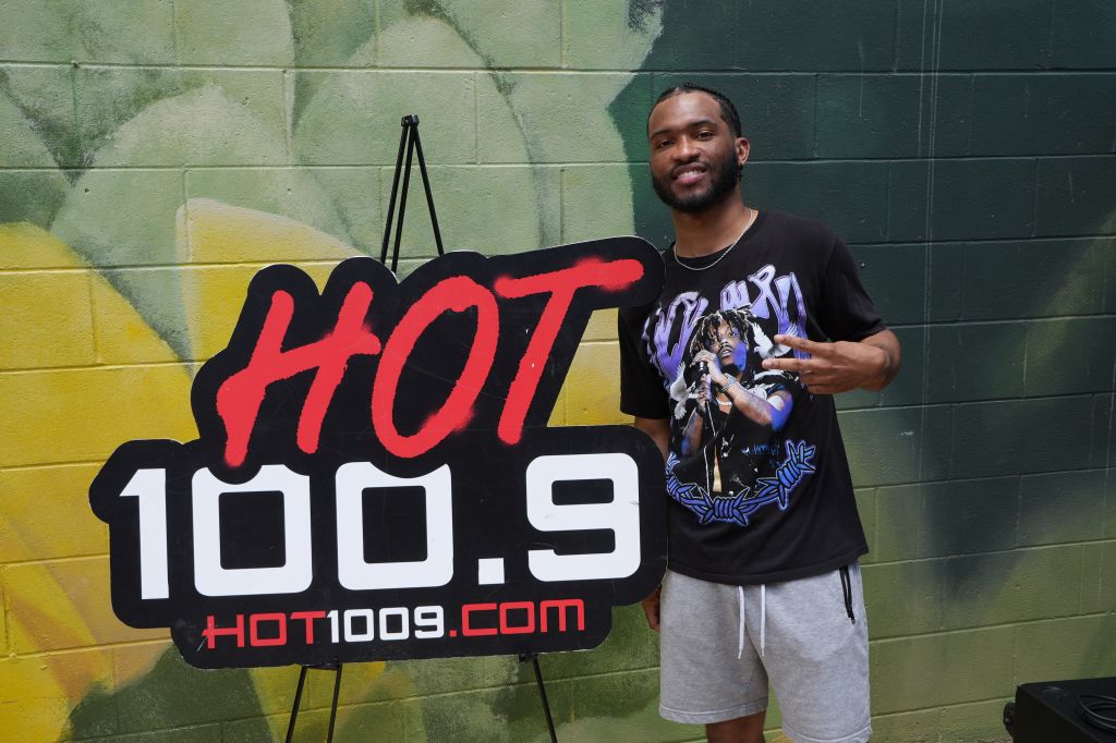 A smiling man standing in front of a large "HOT 100.9" sign, wearing a t-shirt with a graphic design.