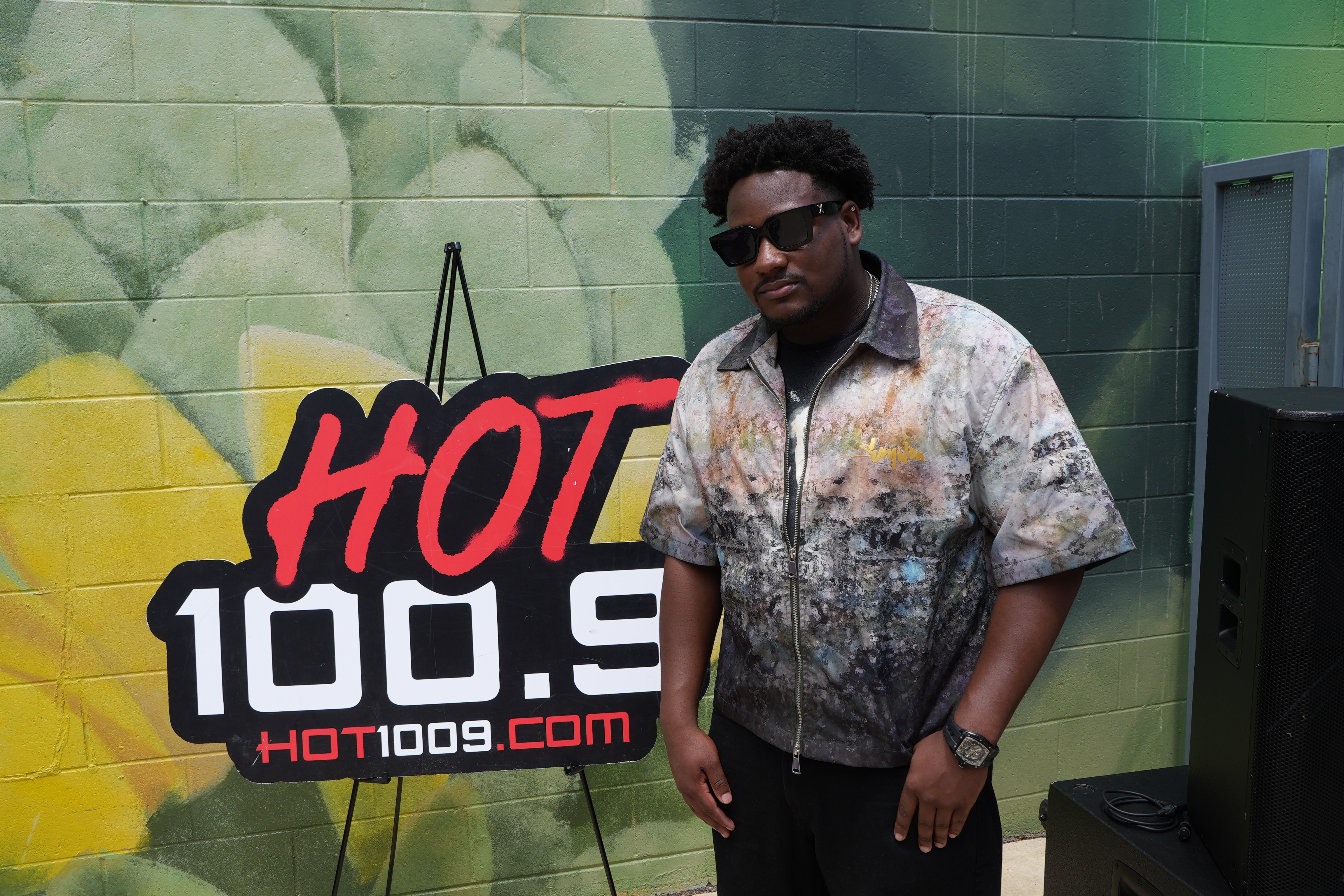A person wearing sunglasses standing in front of a large "HOT 100.9" sign on a green wall.
