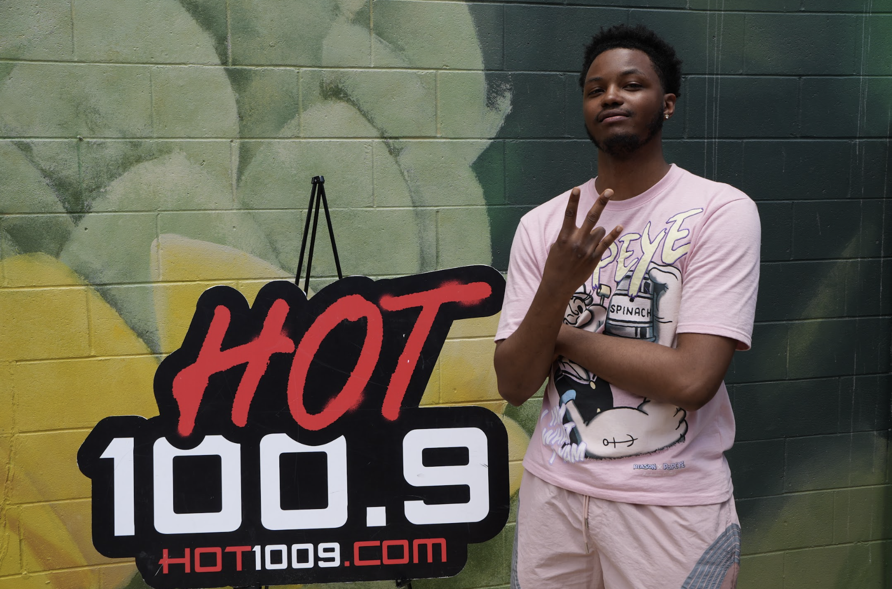 A young man standing in front of a wall with the "HOT 100.9" radio station logo, making a peace sign gesture.