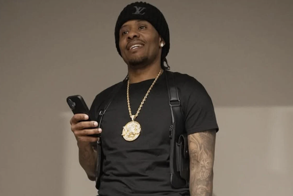 A man wearing a black beanie and gold chain necklace, holding a mobile phone and smiling.