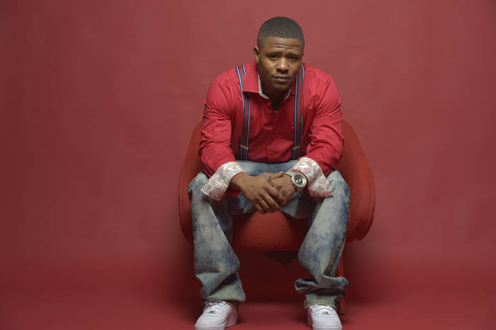 A young Black man wearing a red jacket and jeans sits on a red chair against a red background, looking directly at the camera with a serious expression.