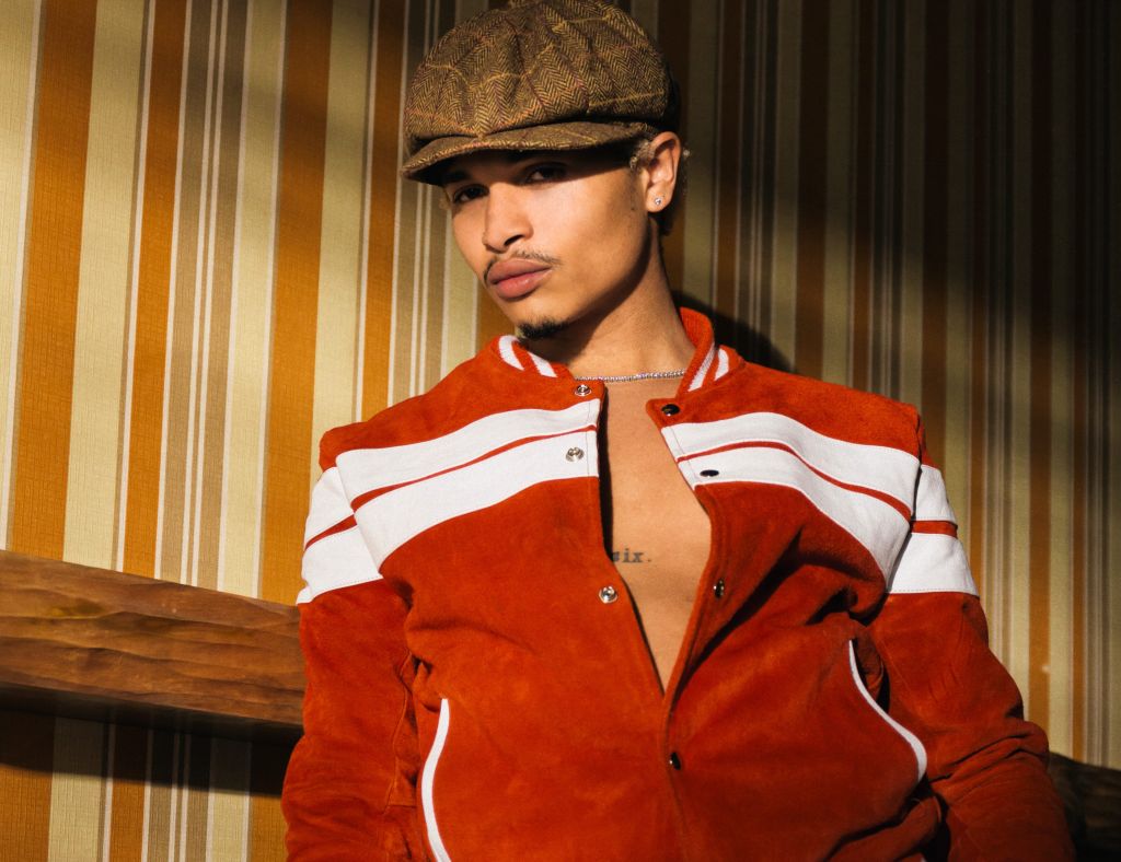 A person wearing a red and white striped jacket and a brown newsboy cap standing in front of a wooden wall.