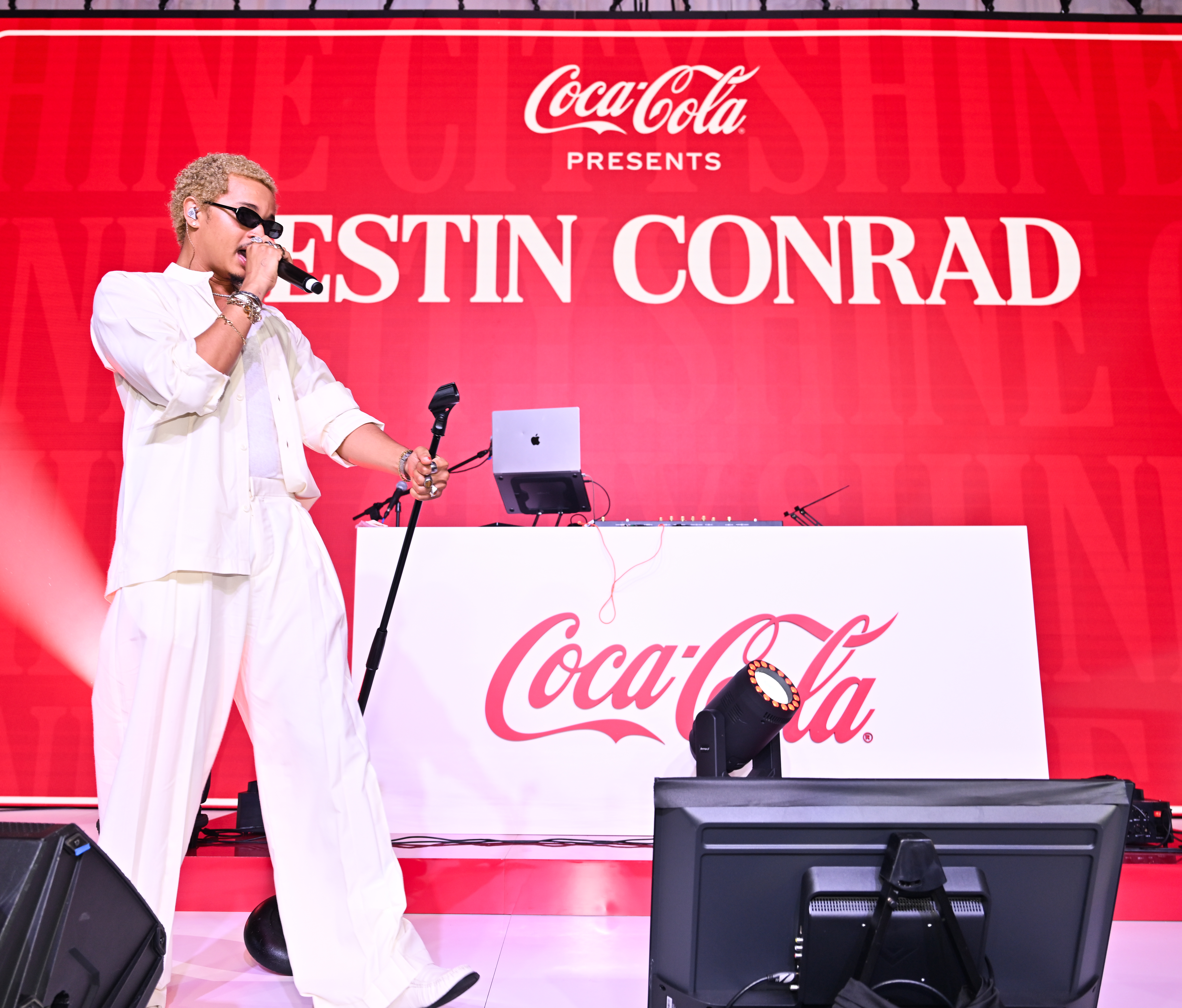 Coca-Cola presents Estin Conrad performing on stage with a laptop and microphone, against a large red Coca-Cola backdrop.