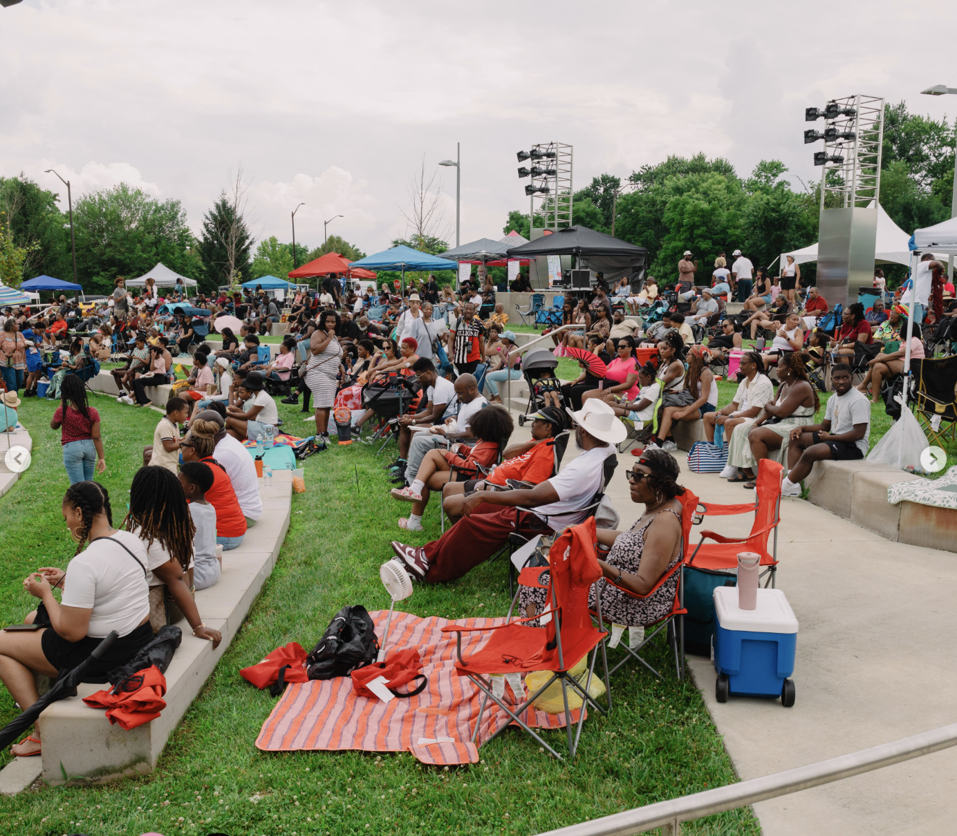 Crowded outdoor event with people sitting on the grass, tents, and a stage with lighting equipment. Colorful umbrellas and chairs dot the grassy area.