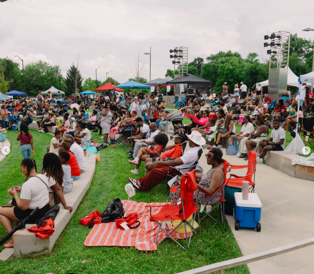 Crowded outdoor event with people sitting on the grass, tents, and a stage with lighting equipment. Colorful umbrellas and chairs dot the grassy area.