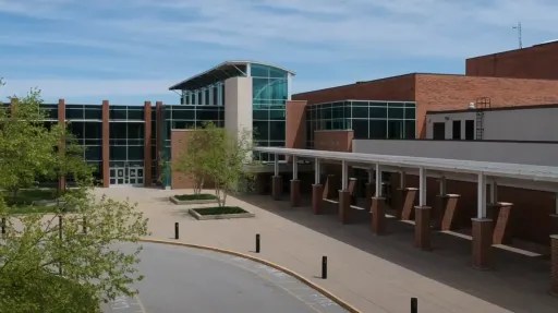 A large brick and glass building with a covered walkway in front, surrounded by trees and a paved area.