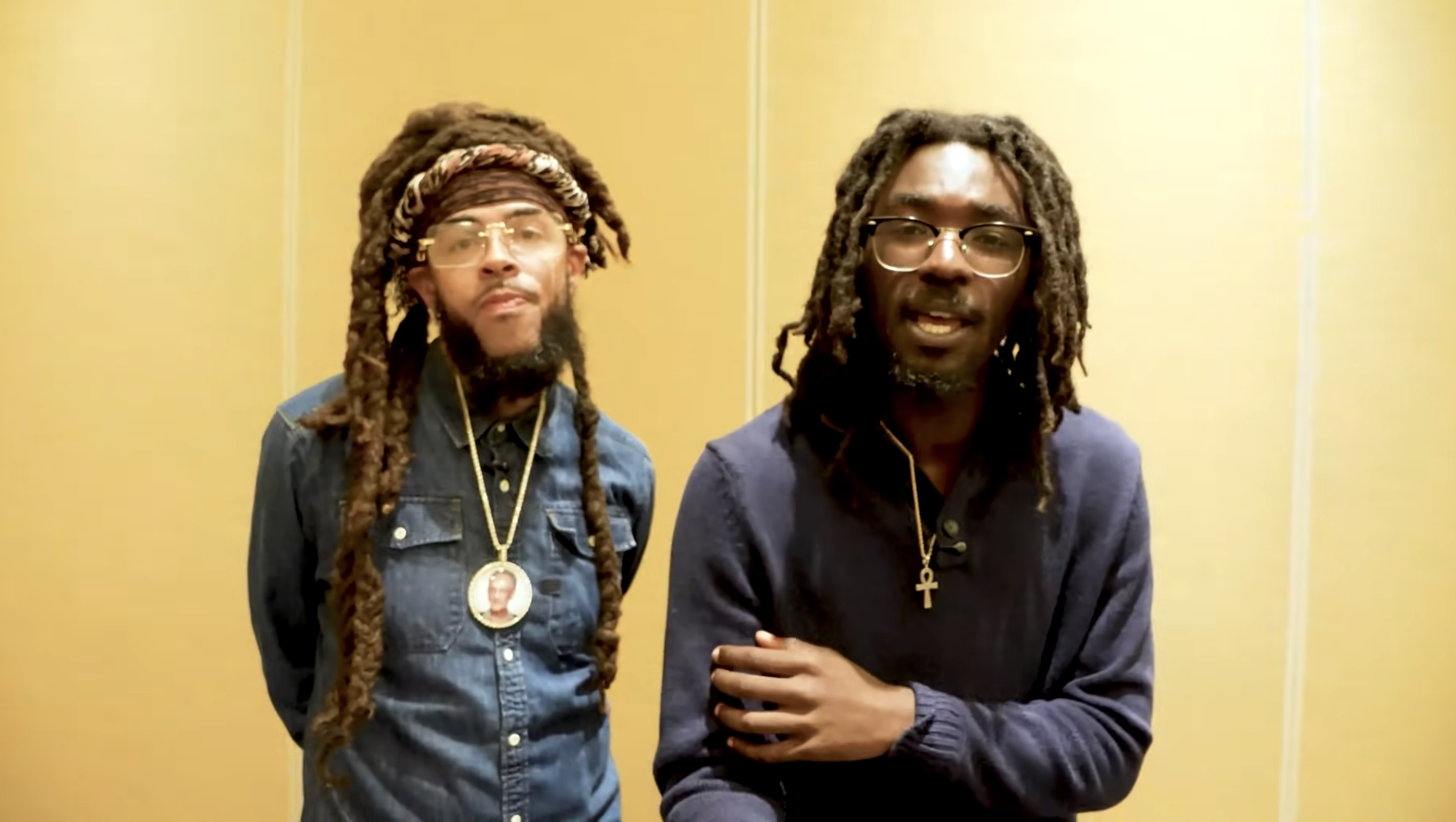 Two men with dreadlocks wearing casual clothing and accessories, standing together in a room with a yellow wall.