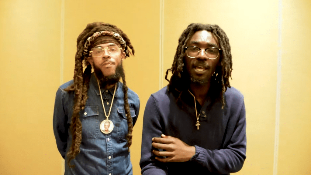 Two men with dreadlocks wearing casual clothing and accessories, standing together in a room with a yellow wall.