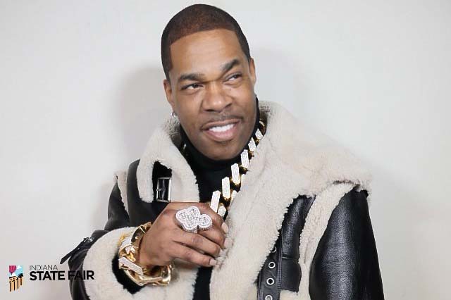 A smiling man wearing a fur-trimmed coat and holding a microphone, standing in front of a backdrop with the Indiana State Fair logo.